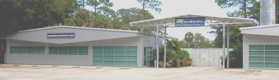 A wide exterior view of a large industrial warehouse or garage building with multiple green-tinted garage-style doors, a covered entryway, trees behind it, and a paved forecourt. A blue sign is visible above the entrance.