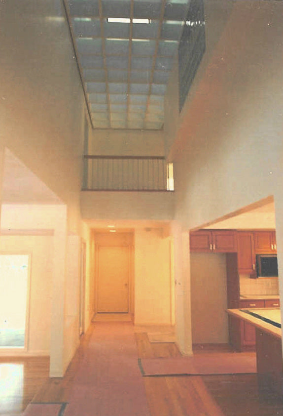 Two-story interior atrium of the Bourque Road Residence in Boston, Massachusetts, featuring a skylight ceiling, open upper gallery, hardwood floors, and an adjoining contemporary kitchen.
