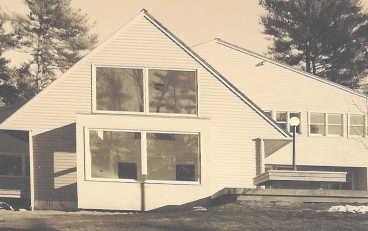 A vintage-style photograph of a modern single-story house with white siding, large angled windows, and a gabled roof, surrounded by trees and an open lawn.