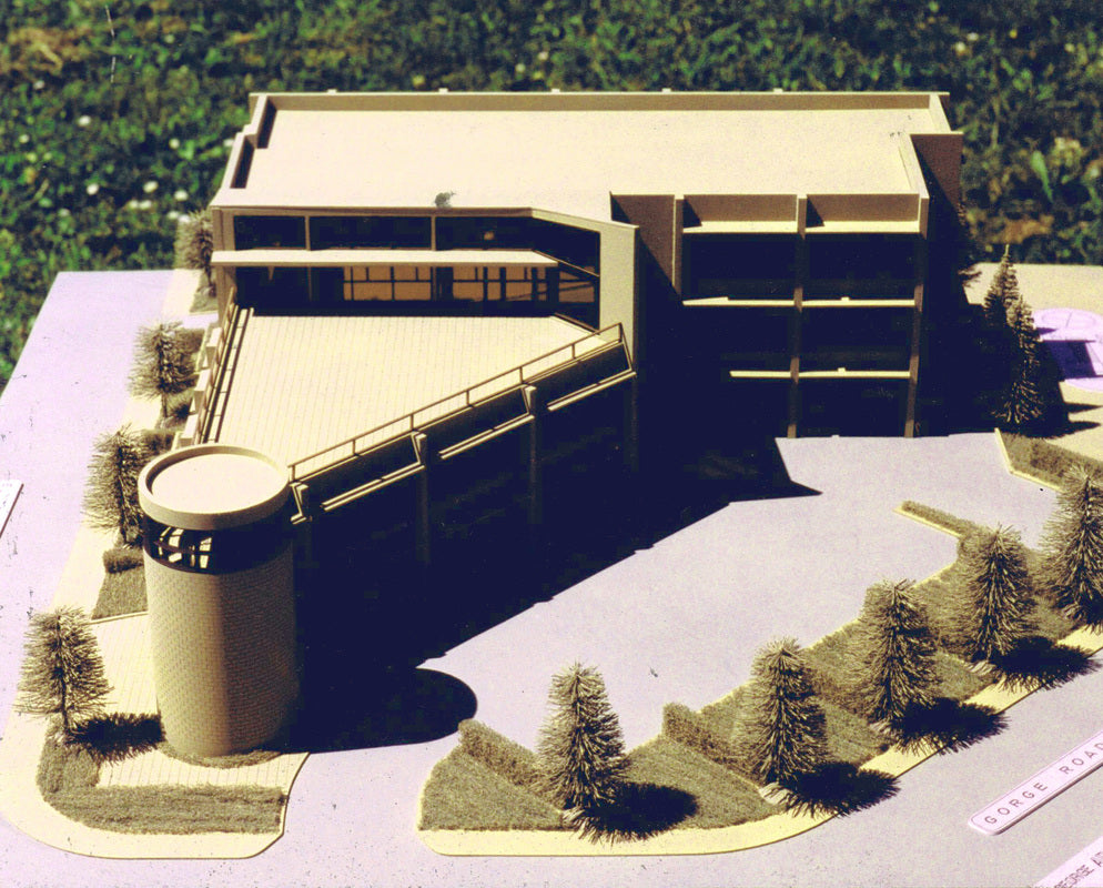 A scale model of a modern multi-story office building with a cylindrical structure, set on a landscaped base with small trees and grass, photographed outdoors.