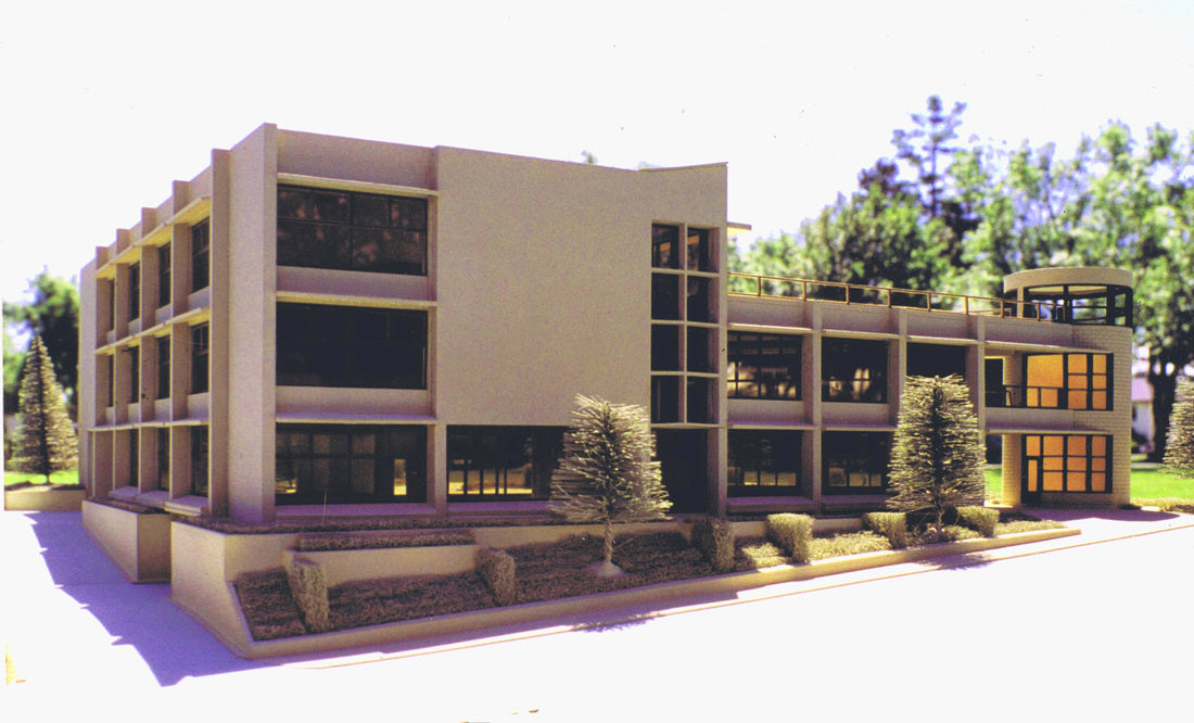 A modern multi-story office building model or rendering with beige exterior walls, large dark windows, landscaped landscaping with shrubs and small trees, and a bright sky background.