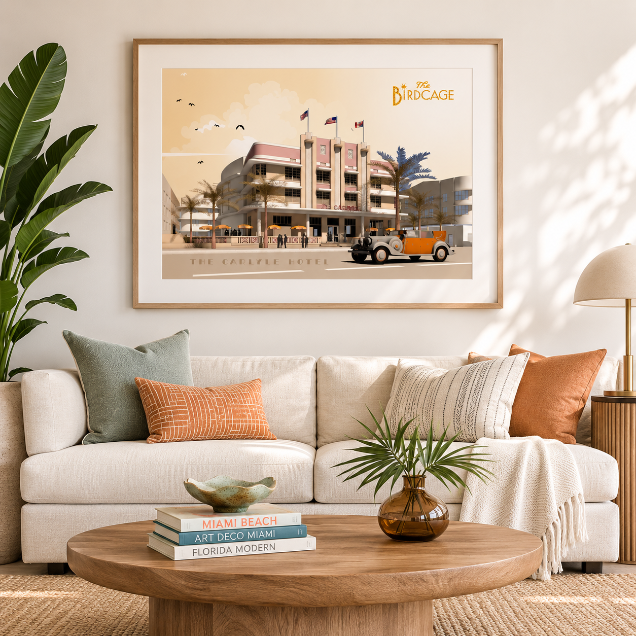 A framed art print titled 'The Bird Cage' hangs on a beige wall above a white sofa in a styled living room. The print depicts a retro mid-century modern scene with a building, palm trees, and a vintage orange car.