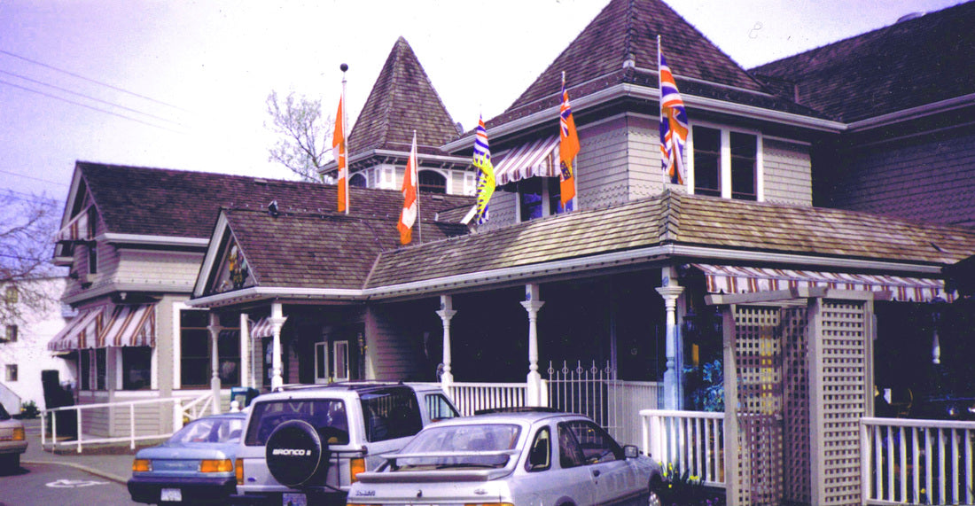 A vintage photograph of a large Victorian-style house with multiple flags on the roof, parked cars in front, and a street scene under a pale sky.
