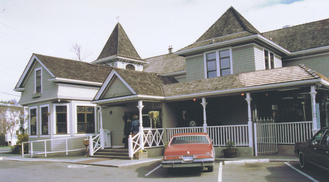 A large historic-style house with a wraparound porch, white railings, gray siding, and brown shingled roofs, with an orange car parked in front on a paved lot.