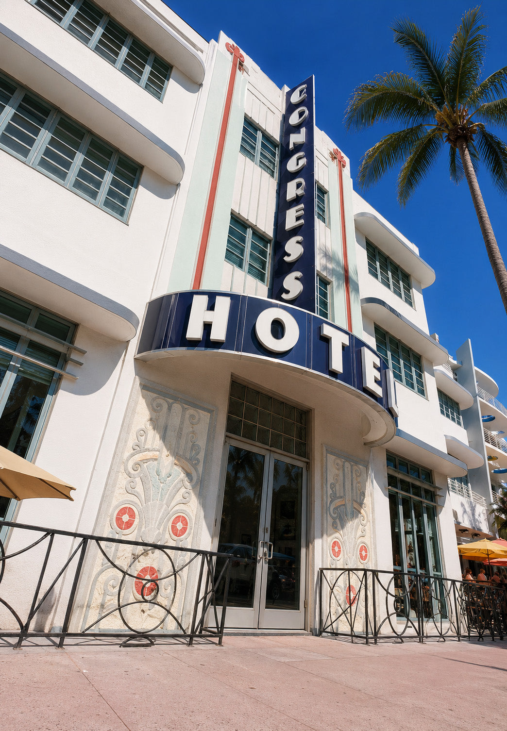 Art Deco style Colgress Hotel facade with a large blue and white marquee sign, white building, palm tree, and blue sky.