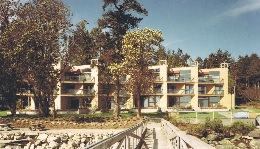 A vintage-style multi-story apartment building with beige walls and large windows, surrounded by tall trees and a landscaped lawn, with a wooden walkway in the foreground.