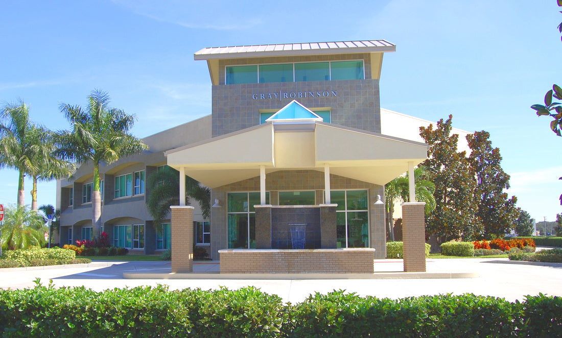 Front entrance of the GrayRobinson office building in Melbourne, Florida, featuring a covered porte-cochère, central fountain, three-story entry tower, and contemporary architecture surrounded by palm trees and landscaped grounds.